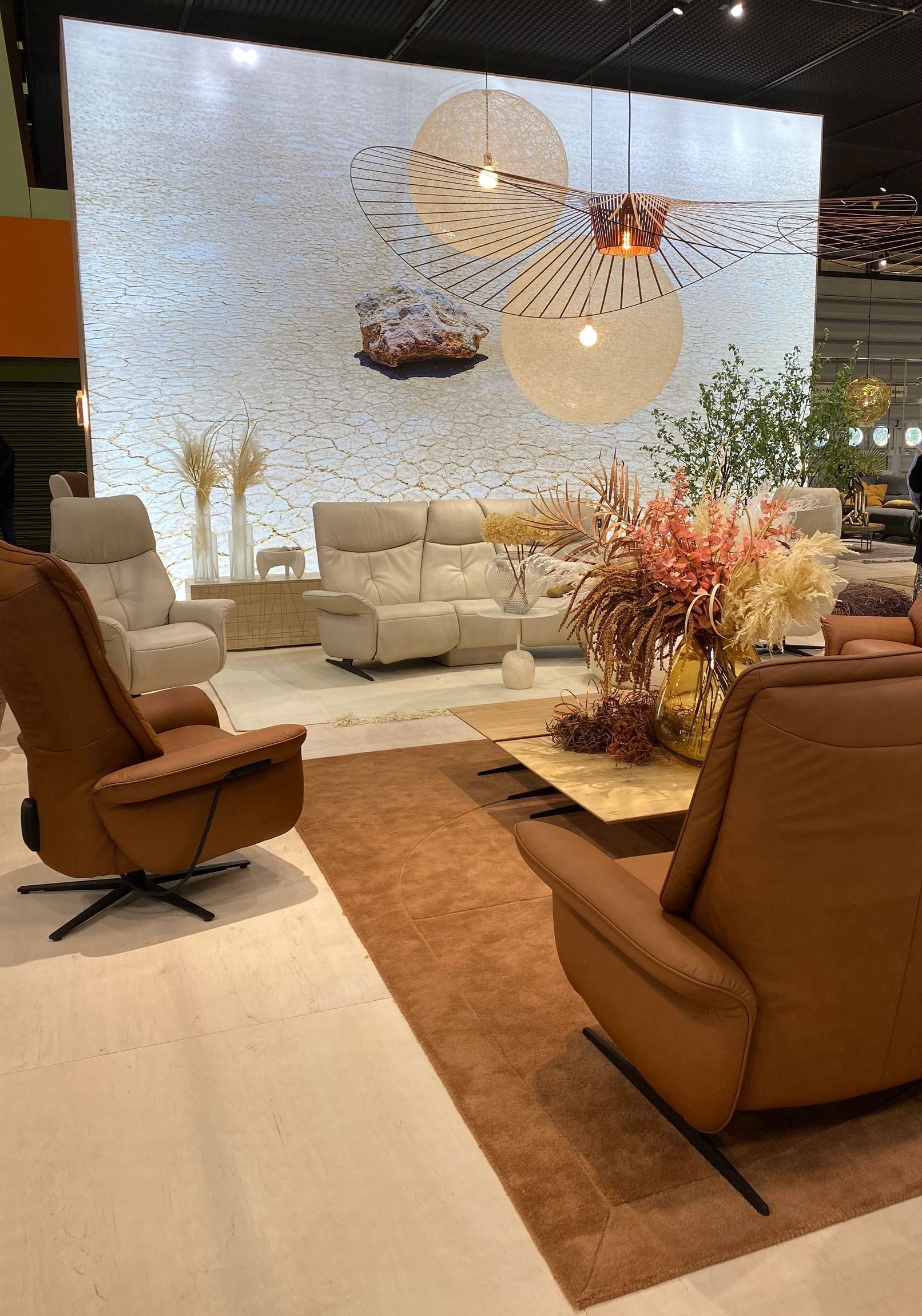 living room display with luxury brown leather recliner swivel chairs and a white leather sofa system and large low level coffee table with extensive floral display and bespoke lighting unit overhead at the January Furniture Show 2024, NEC, Birmingham