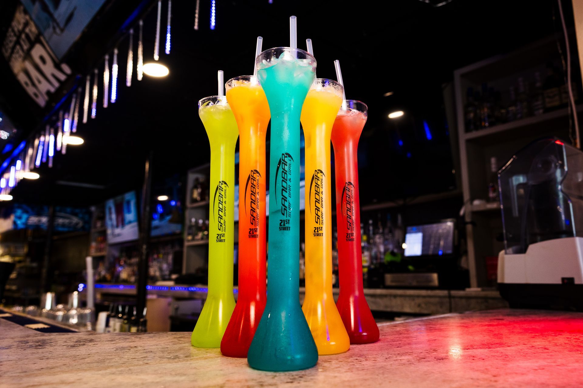 A row of tall glasses filled with different colored drinks on a bar.