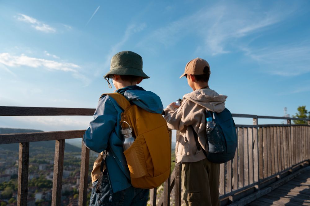 Duas crianças com mochilas contemplam a vista panorâmica de uma ponte de madeira, sob um céu azul.