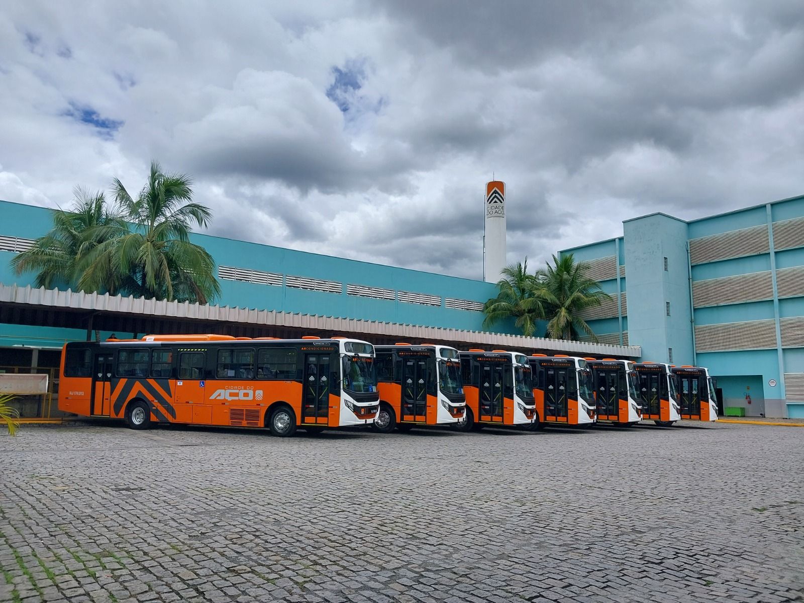 Três ônibus de passageiros brancos estacionados em um terreno pavimentado, com uma encosta verde ao fundo.