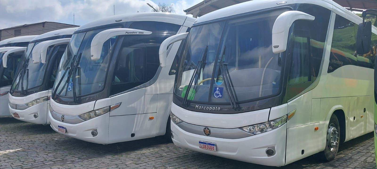 Três ônibus brancos estacionados em fila num terreno sob um céu nublado.