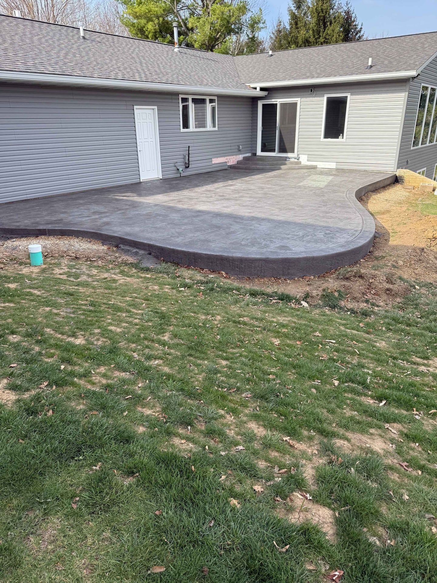 Concrete patio with curved edge, gray siding house. Green grass, blue pipe, and a door are visible.