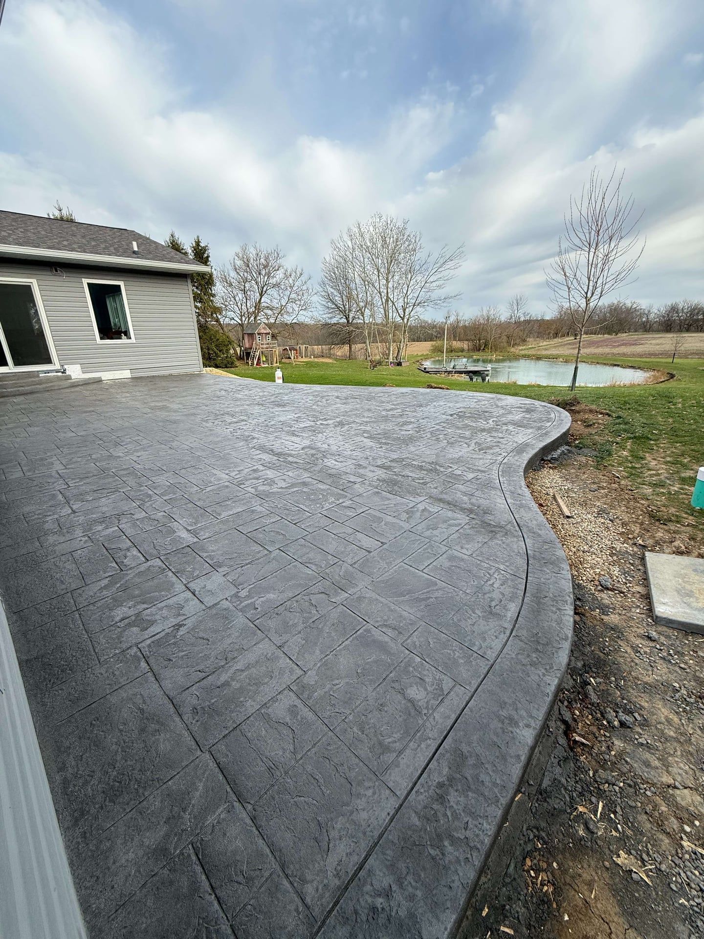 Gray concrete patio with a curved edge, overlooking a field and small pond.
