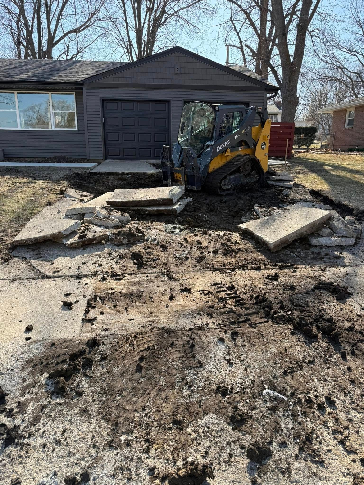 A small construction vehicle demolishes a concrete driveway in front of a gray house and garage.