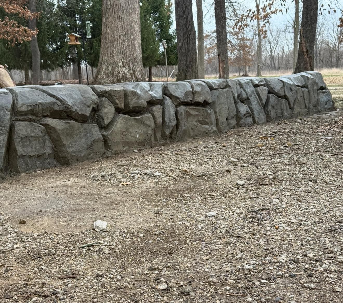 Stone wall in an outdoor setting, with trees in the background and a dirt ground.