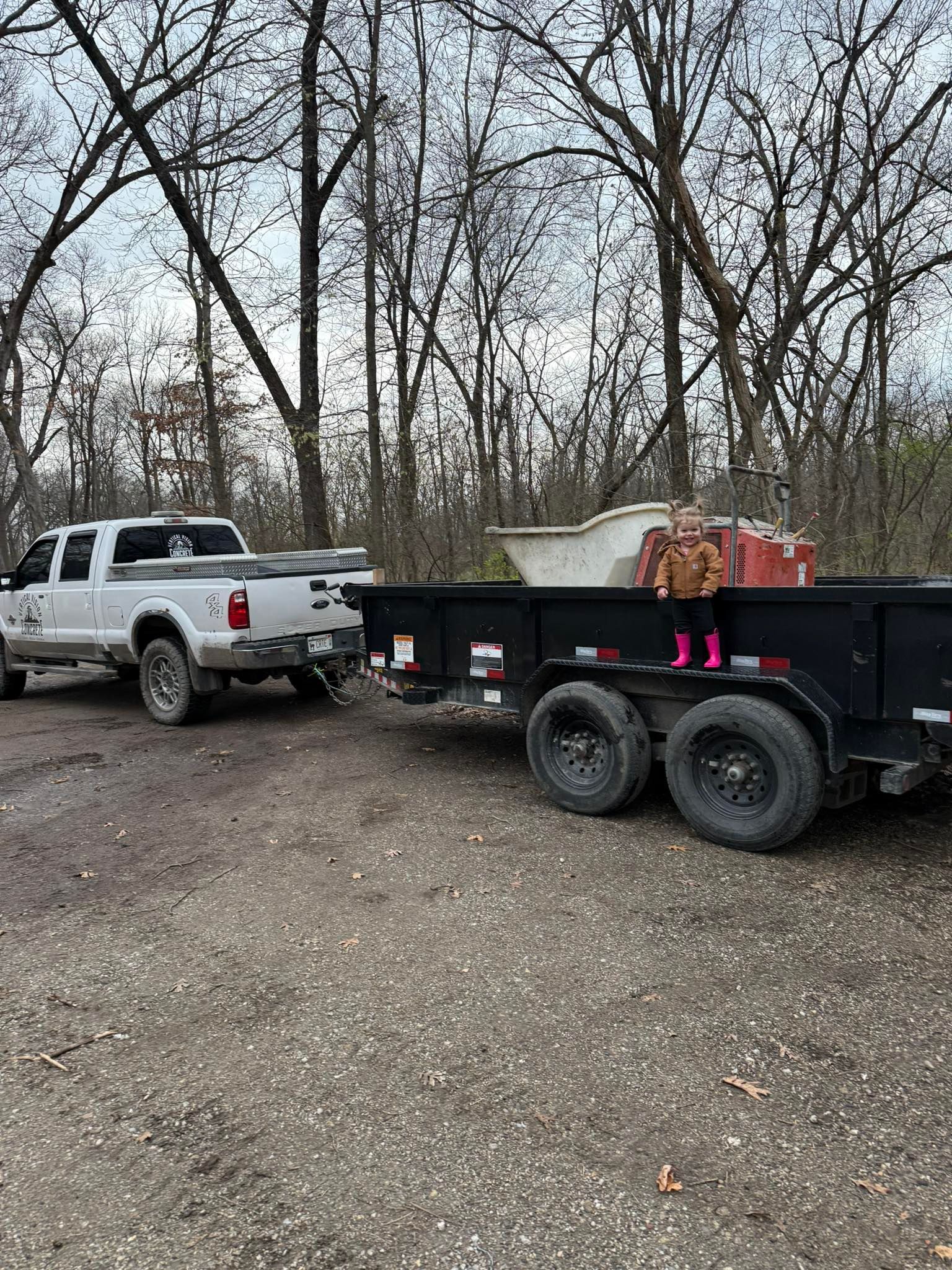 White pickup truck towing a black trailer loaded with a bathtub and debris on a gravel road.