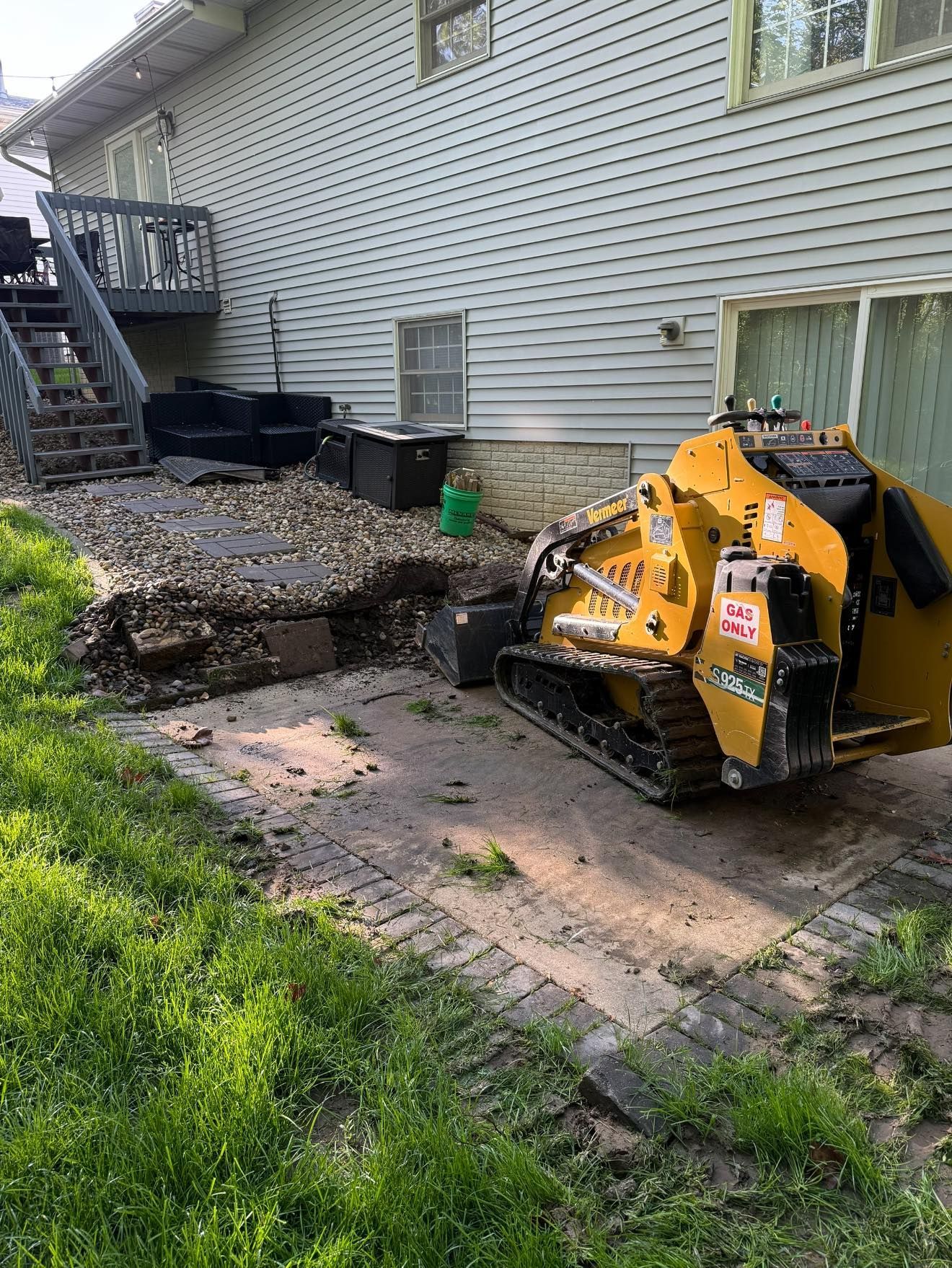 Yellow skid steer removing material from a backyard, near a house with stairs and a patio.