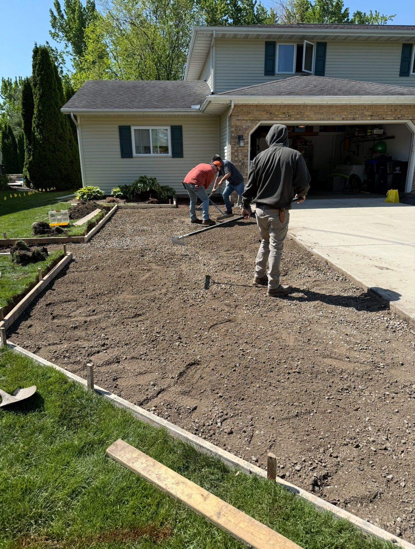 Men working on a driveway project. One man is walking, two are working with tools. House and lawn in background.