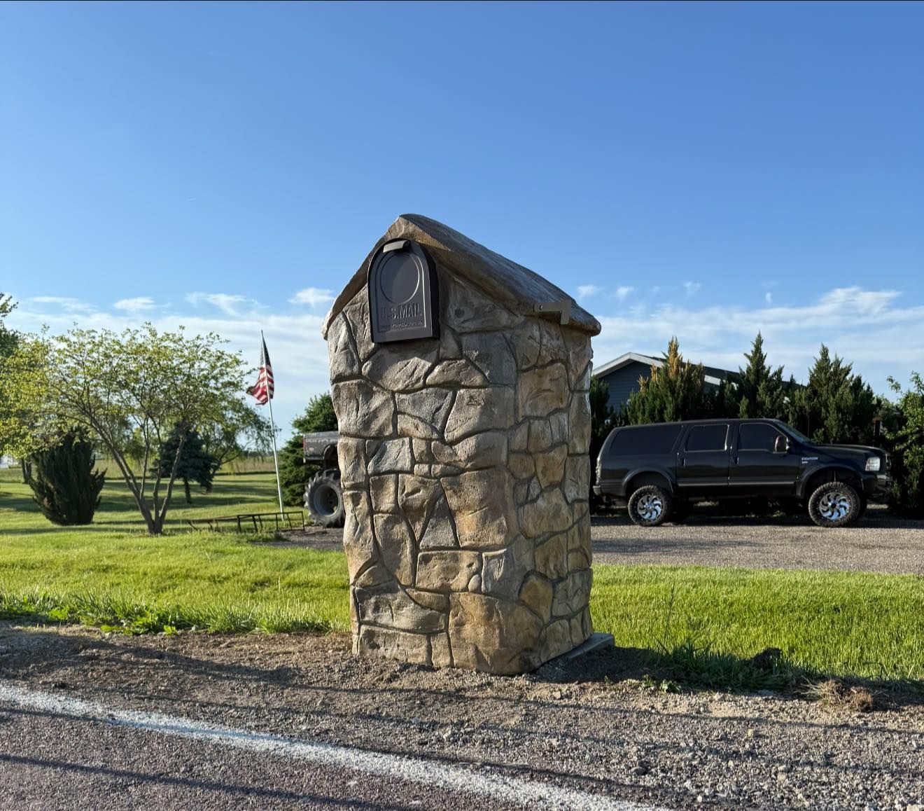 Stone mailbox with a plaque, next to a road, with a dark SUV parked behind it.
