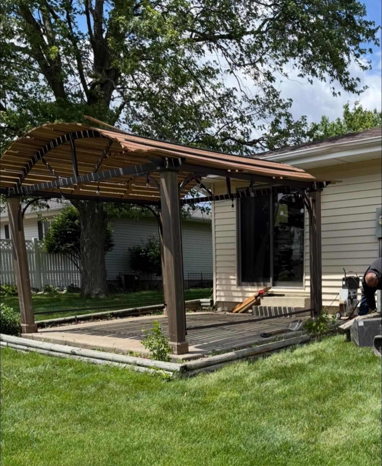 Pergola over a patio with string lights, next to a house with grassy lawn.