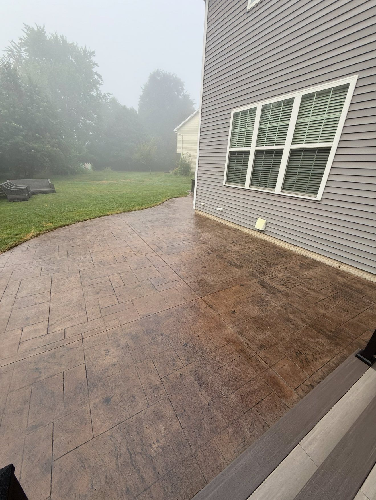 Patio with textured brown concrete leading to a lawn on a foggy day next to a gray house.