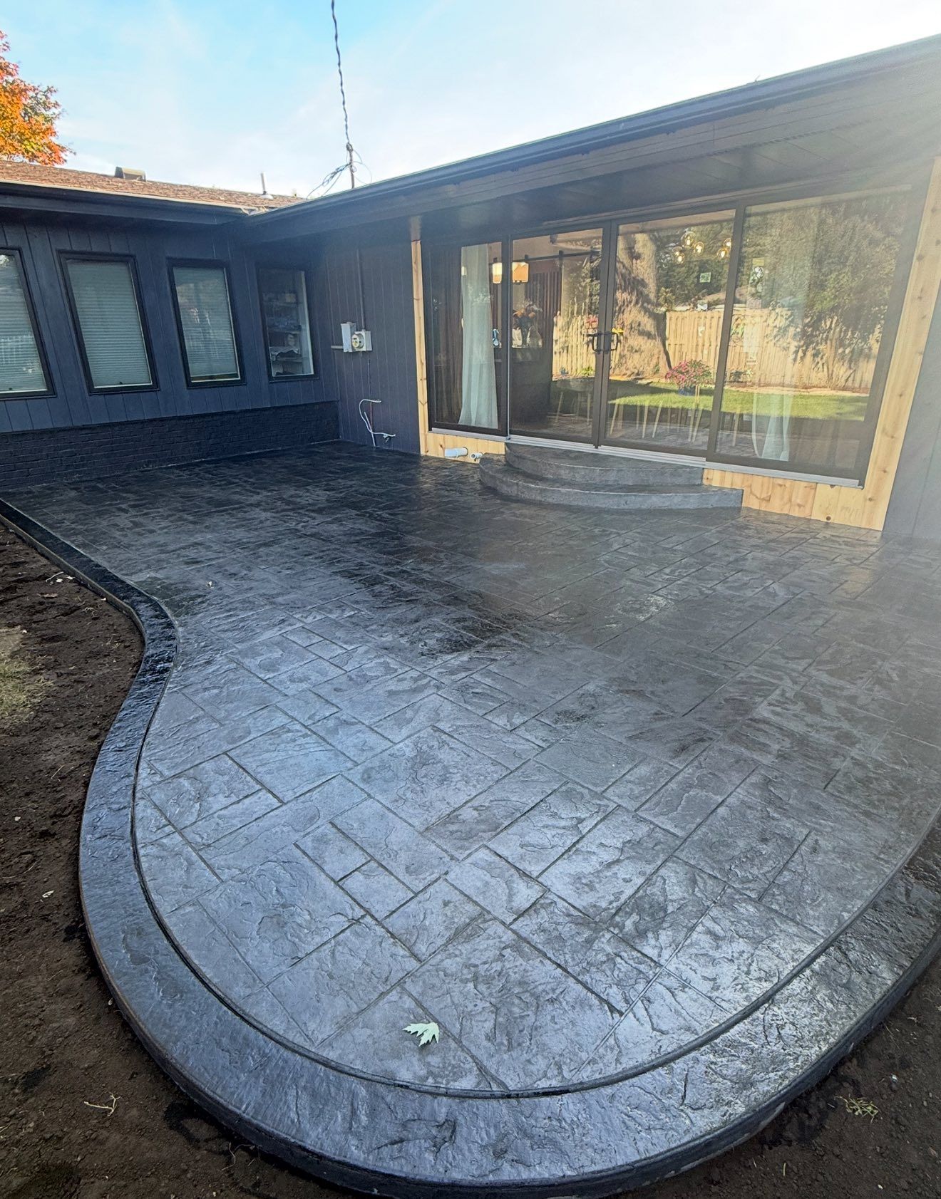 Newly poured, dark gray stamped concrete patio curves along a house with large windows.