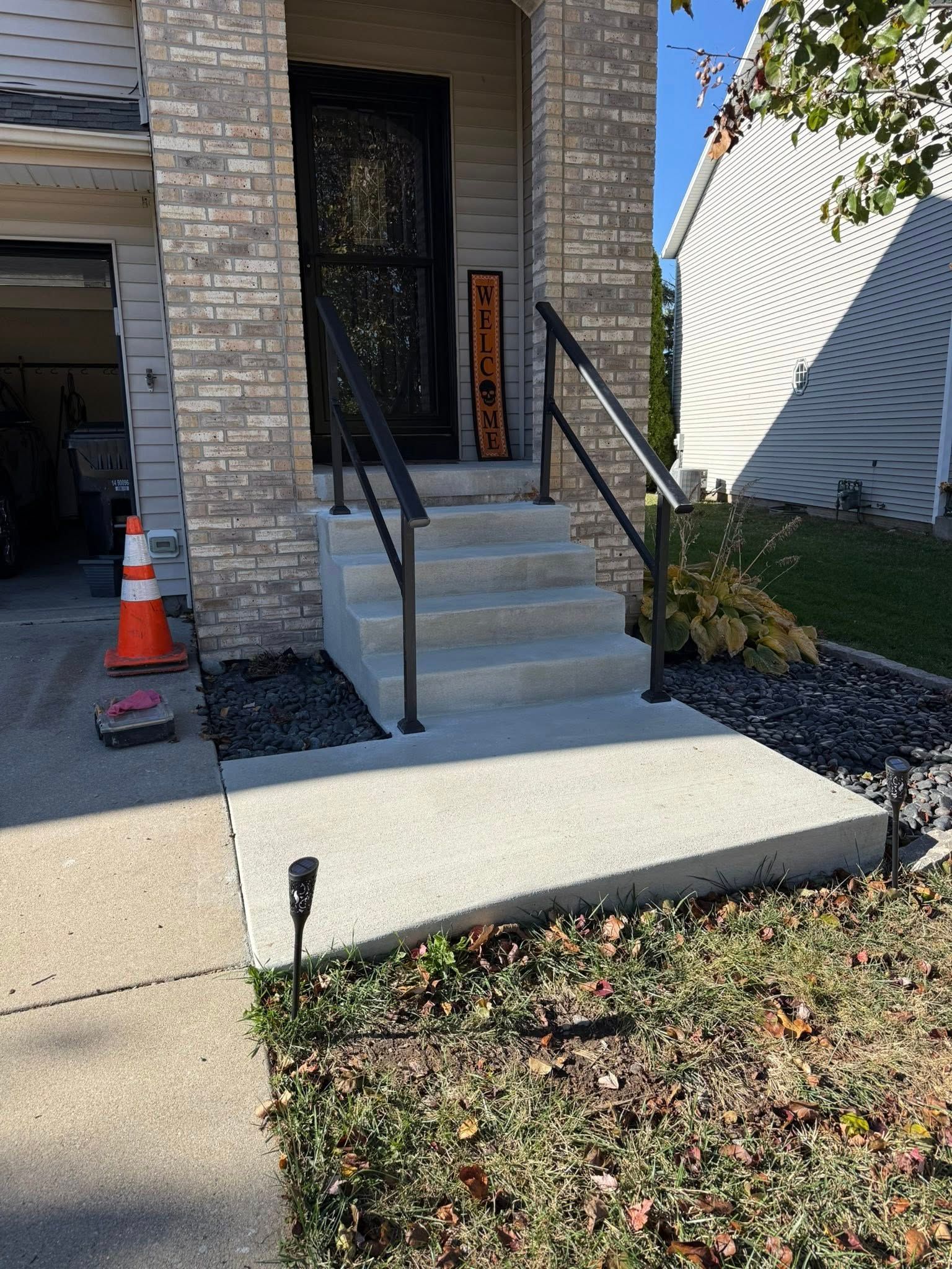 A house entryway with four concrete steps leading to a front door, flanked by black metal handrails and dark landscaping.
