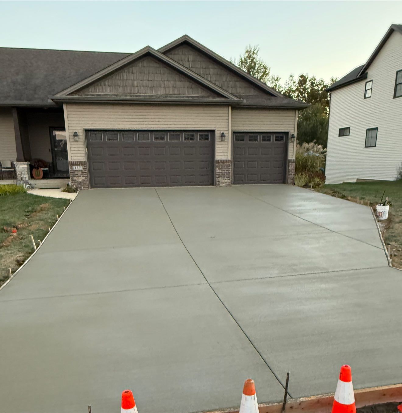 New concrete driveway in front of a tan house with a two-car garage. Orange traffic cones line the edge.