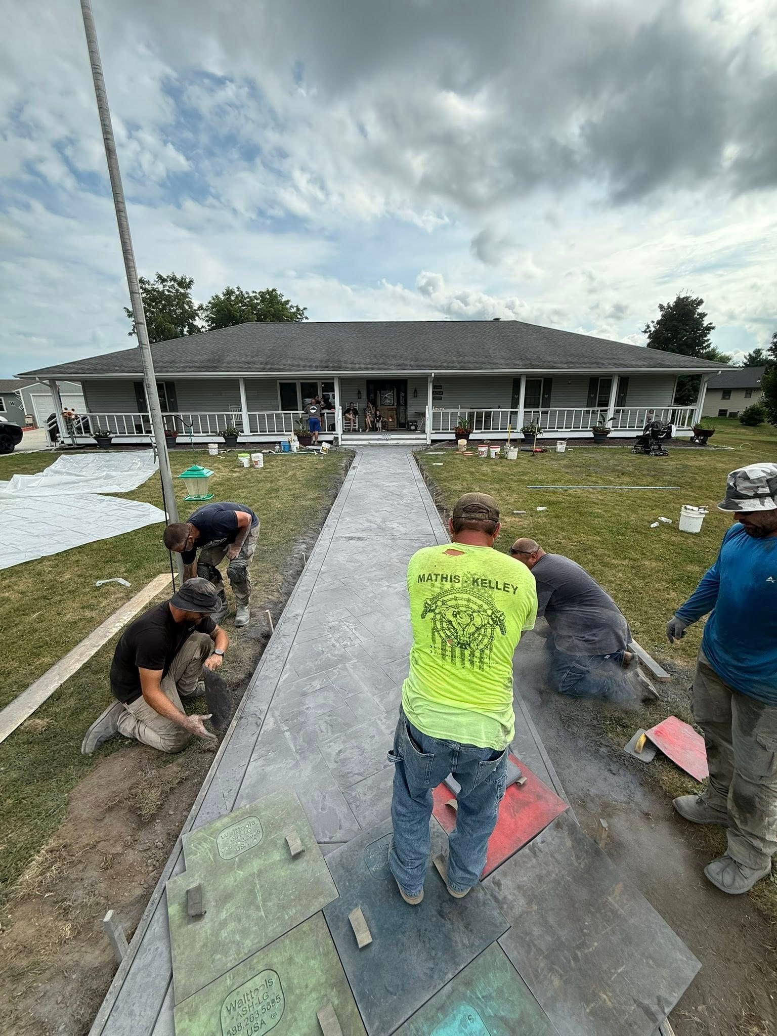 Construction workers laying paving stones for a walkway in front of a gray house with a cloudy sky overhead.