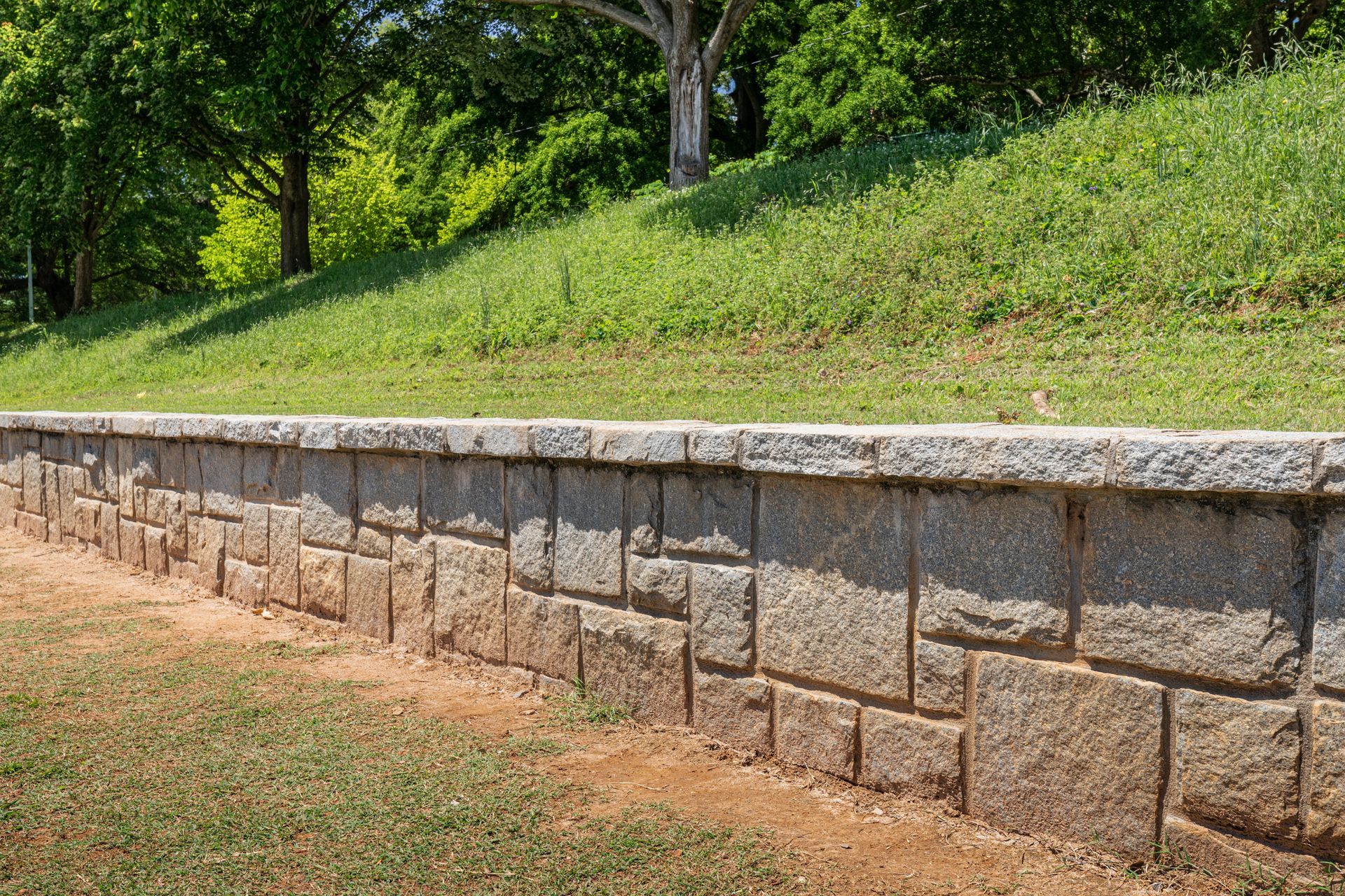Stone retaining wall in front of a grassy hill with trees in the background.