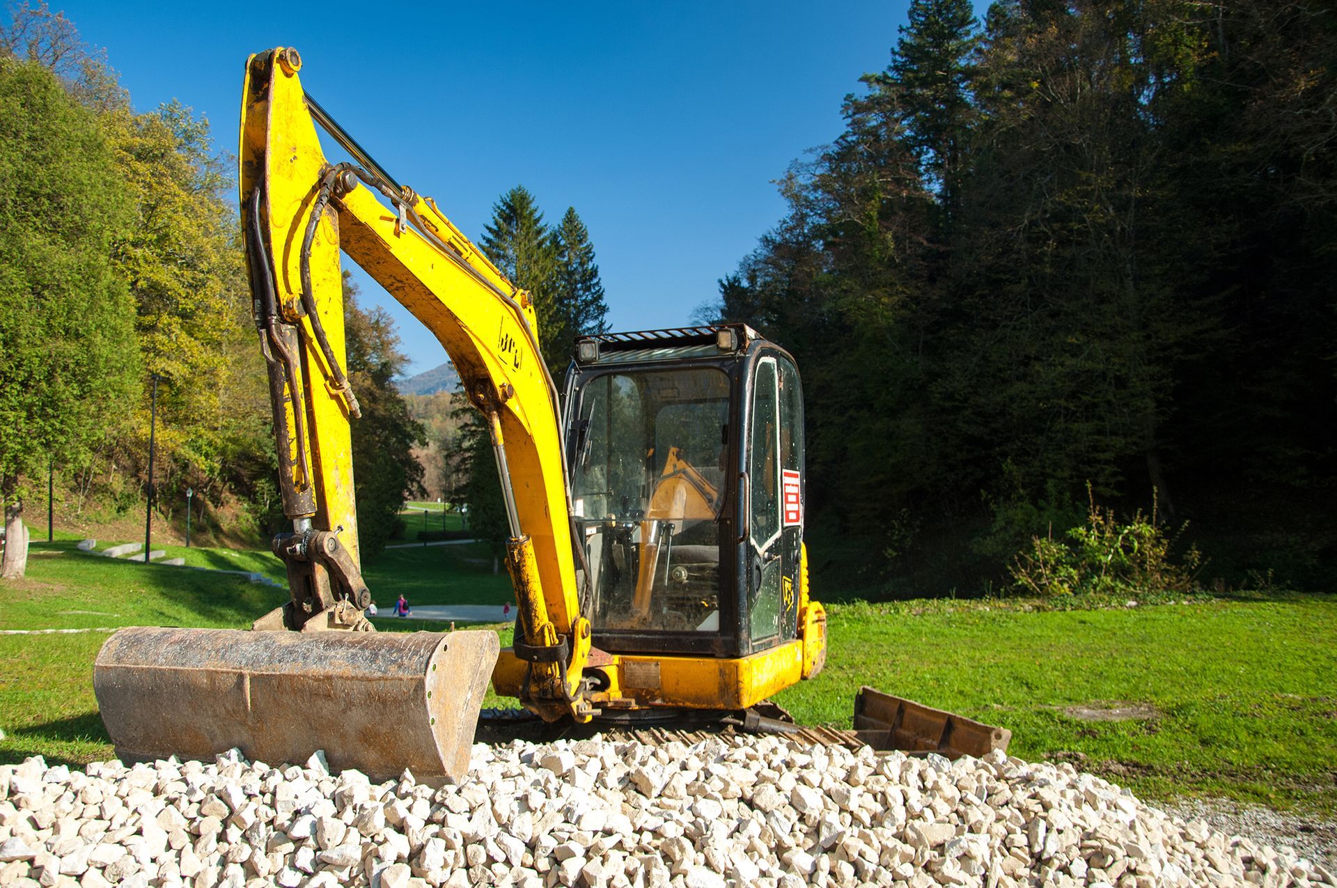 Yellow excavator on a pile of gravel, with a green lawn and trees in the background under a blue sky.