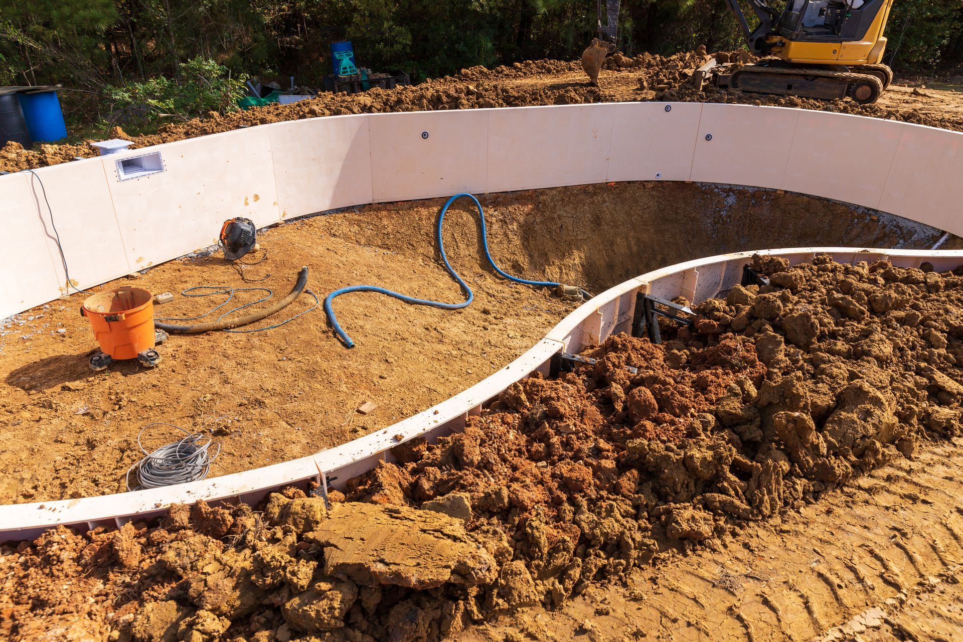 Pool construction site: excavated ground, curved walls, pipes, sand, orange bucket.