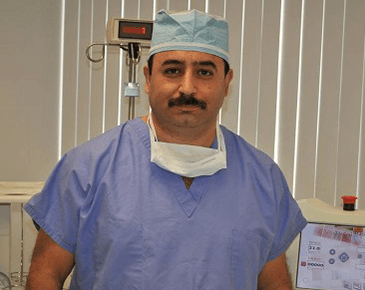 Surgeon in blue scrubs and cap, mask around neck, in a medical setting.