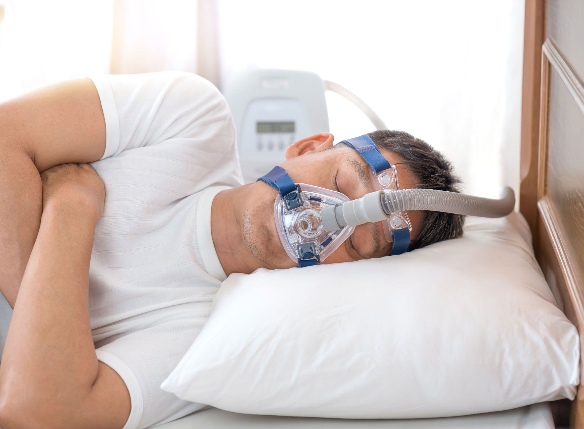 Man wearing a CPAP mask sleeping in bed with a white pillow and machine in the background.