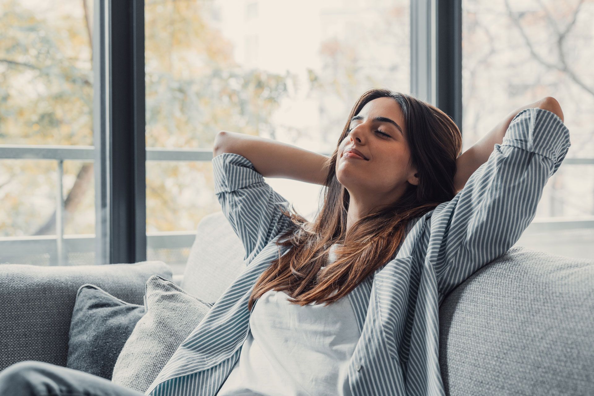 Woman relaxing on a gray sofa, arms behind her head, eyes closed. Sunlight streams through a window.