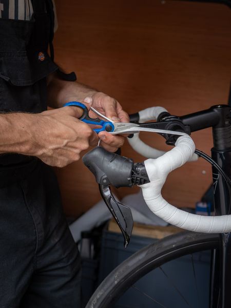 Person cutting white handlebar tape on a black road bike with blue scissors.