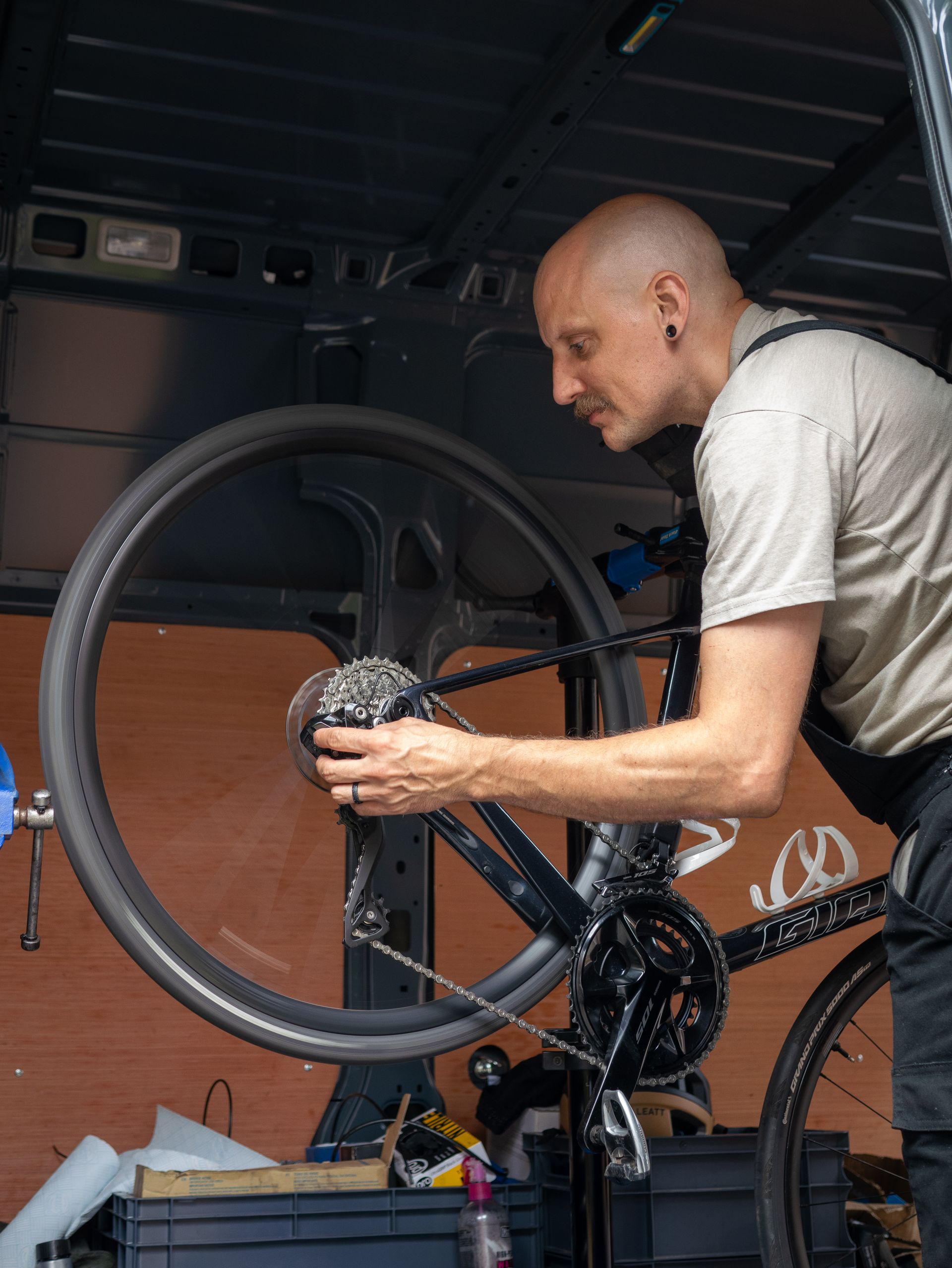 Man fixing a bicycle in a van. Dark bike with silver details, man wears work clothes and has a mustache.