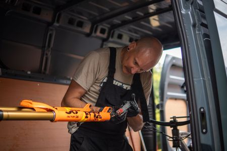 A man working on a mountain bike fork inside a van. He is wearing gloves and overalls. The fork is orange.