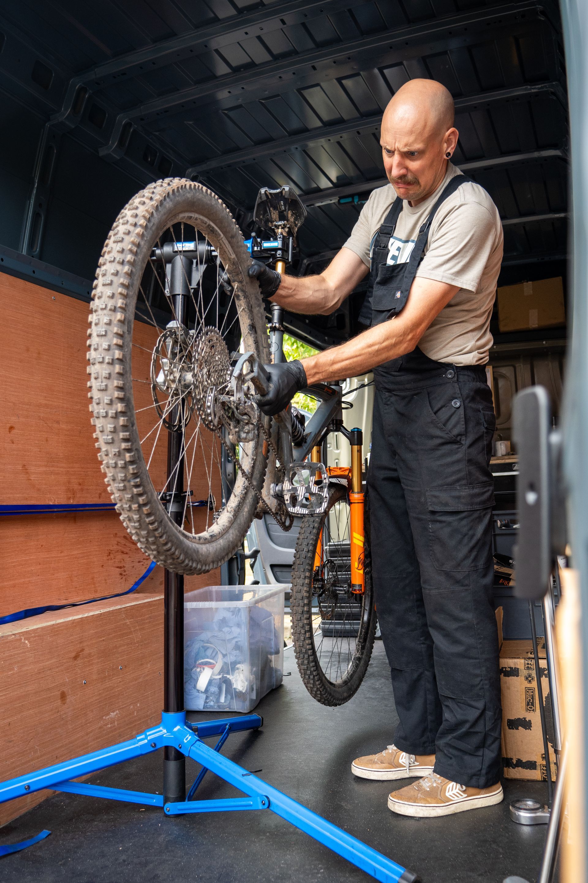 Man working on a teal bicycle, likely for a bike tune-up, outside a van with 