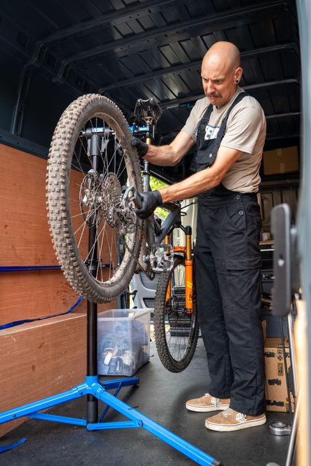 Man in overalls repairs a mountain bike inside a van. He's wearing gloves and focused on the rear wheel.