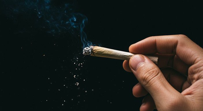 A close-up of a hand holding a burning marijuana joint with ash falling