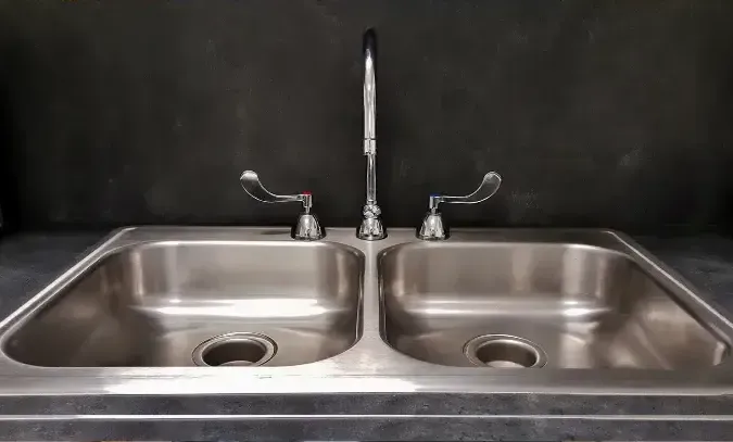 Stainless steel double sink with chrome faucet and two lever handles against a dark backsplash.