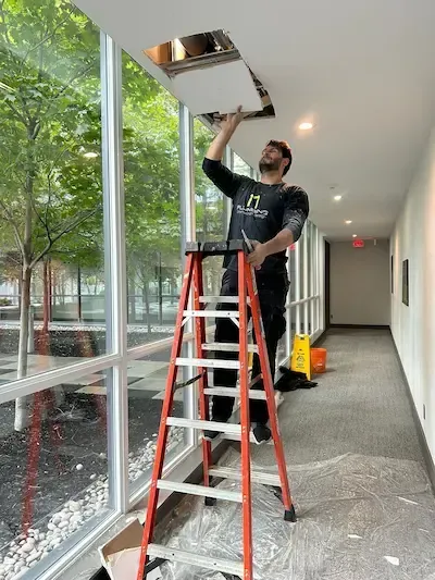 Licensed plumber performing ceiling plumbing repair in Oakville condominium building using ladder and safety equipment.