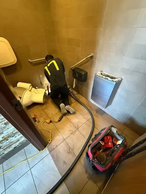 Plumber kneeling with tools and equipment bag while performing toilet snaking service in McDonald’s Oakville restroom.