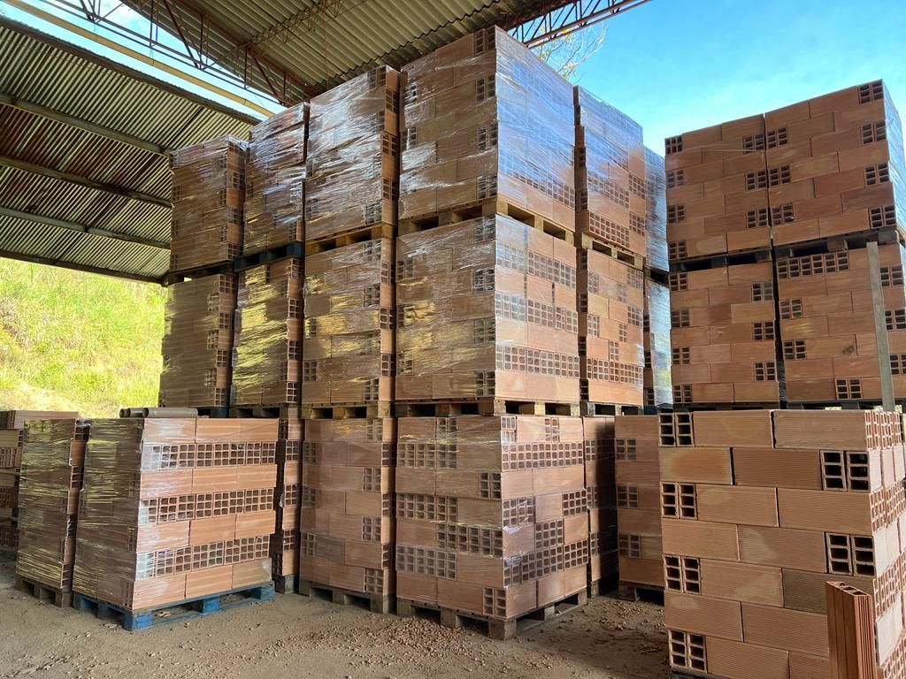 Pilas de ladrillos huecos de arcilla de color marrón rojizo, envueltos en plástico transparente, sobre paletas de madera bajo un techo industrial abierto por los lados.