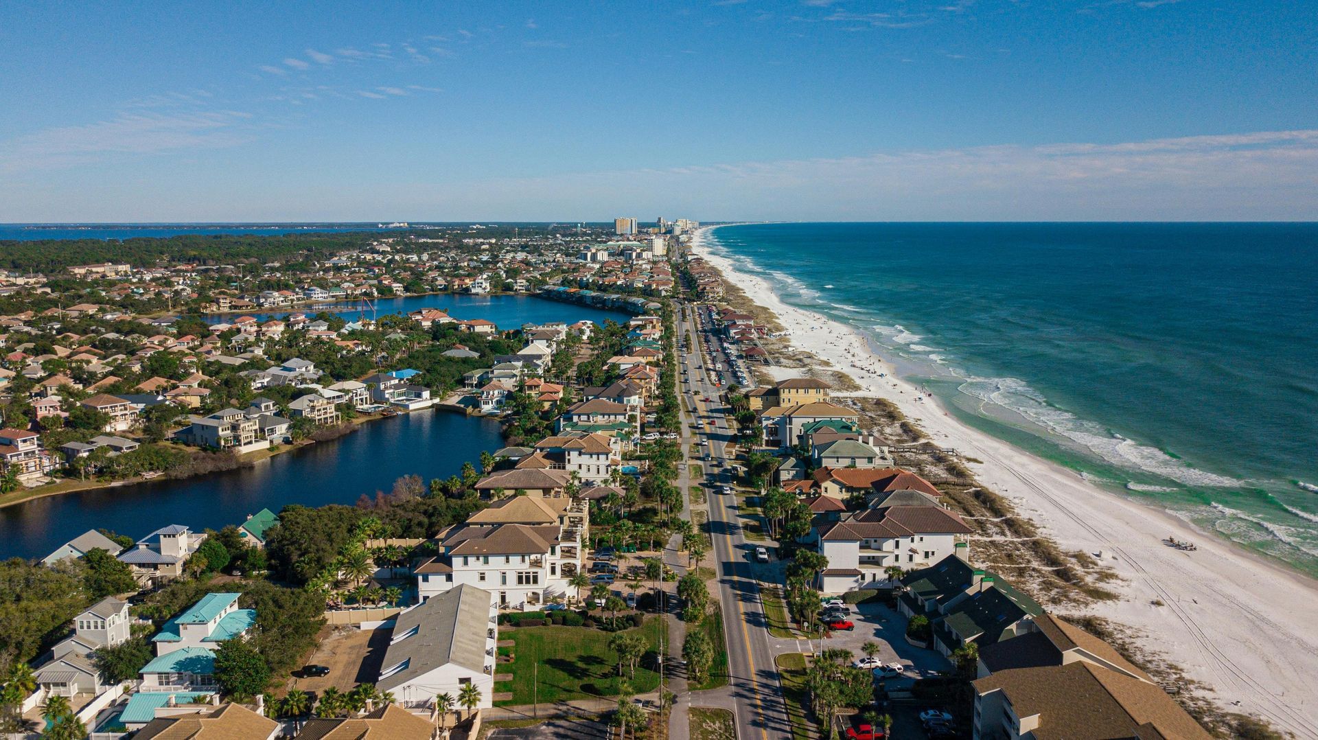 Aerial view of a beach town with white sand, turquoise water, buildings, a road, and a lake on a sunny day.