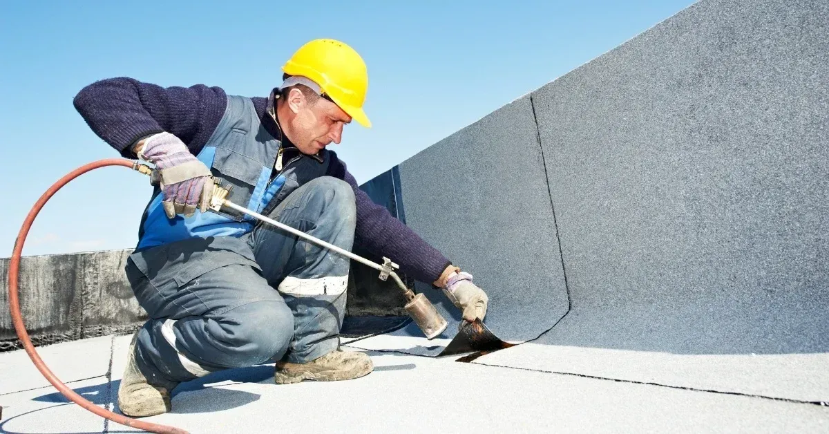 A man is kneeling down on a roof using a torch.