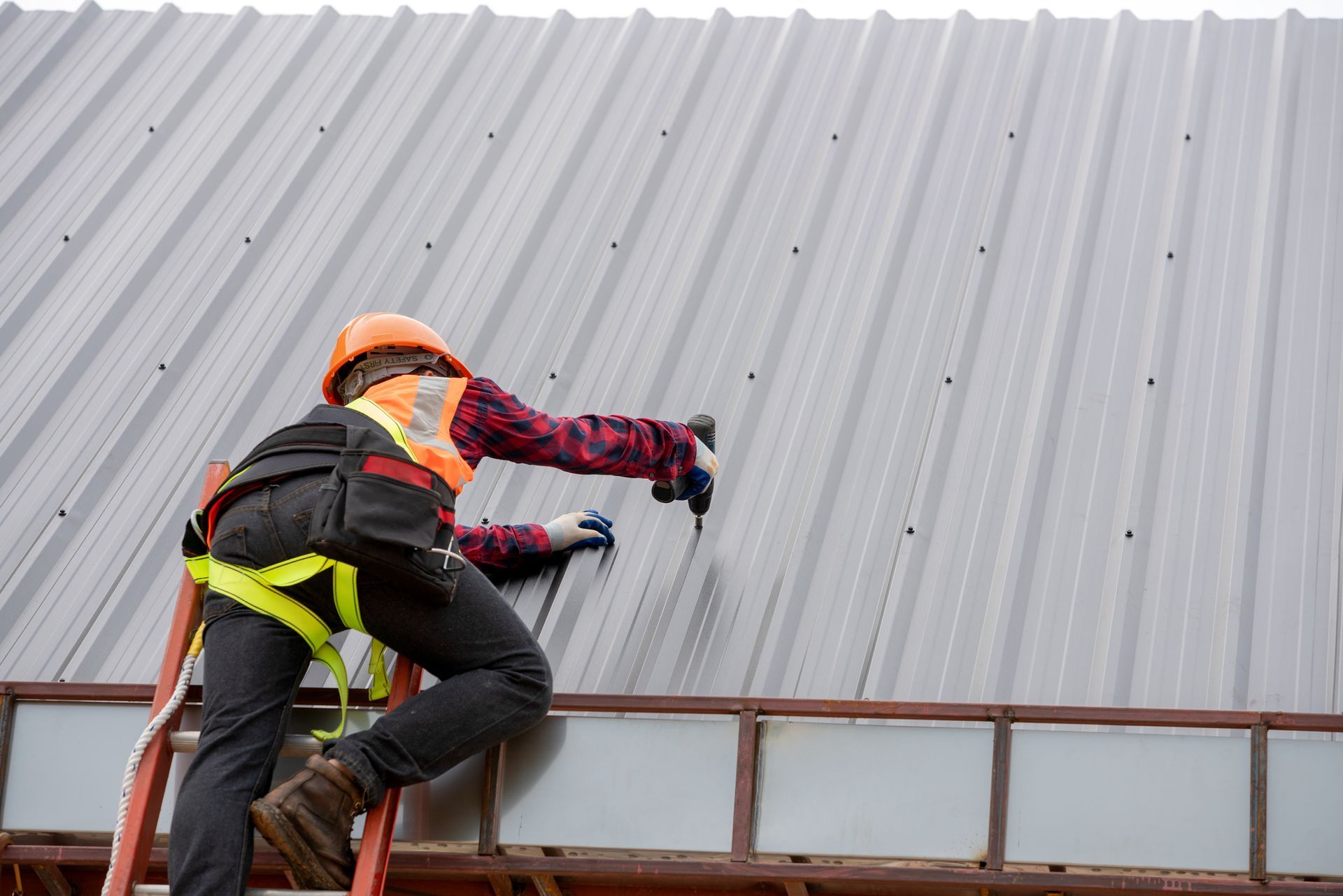 Worker in safety gear installing metal roofing panels while standing on a ladder.