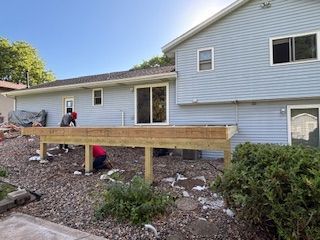 Workers building a wooden deck beside a house on a gravel yard, with one person crouching underneath.