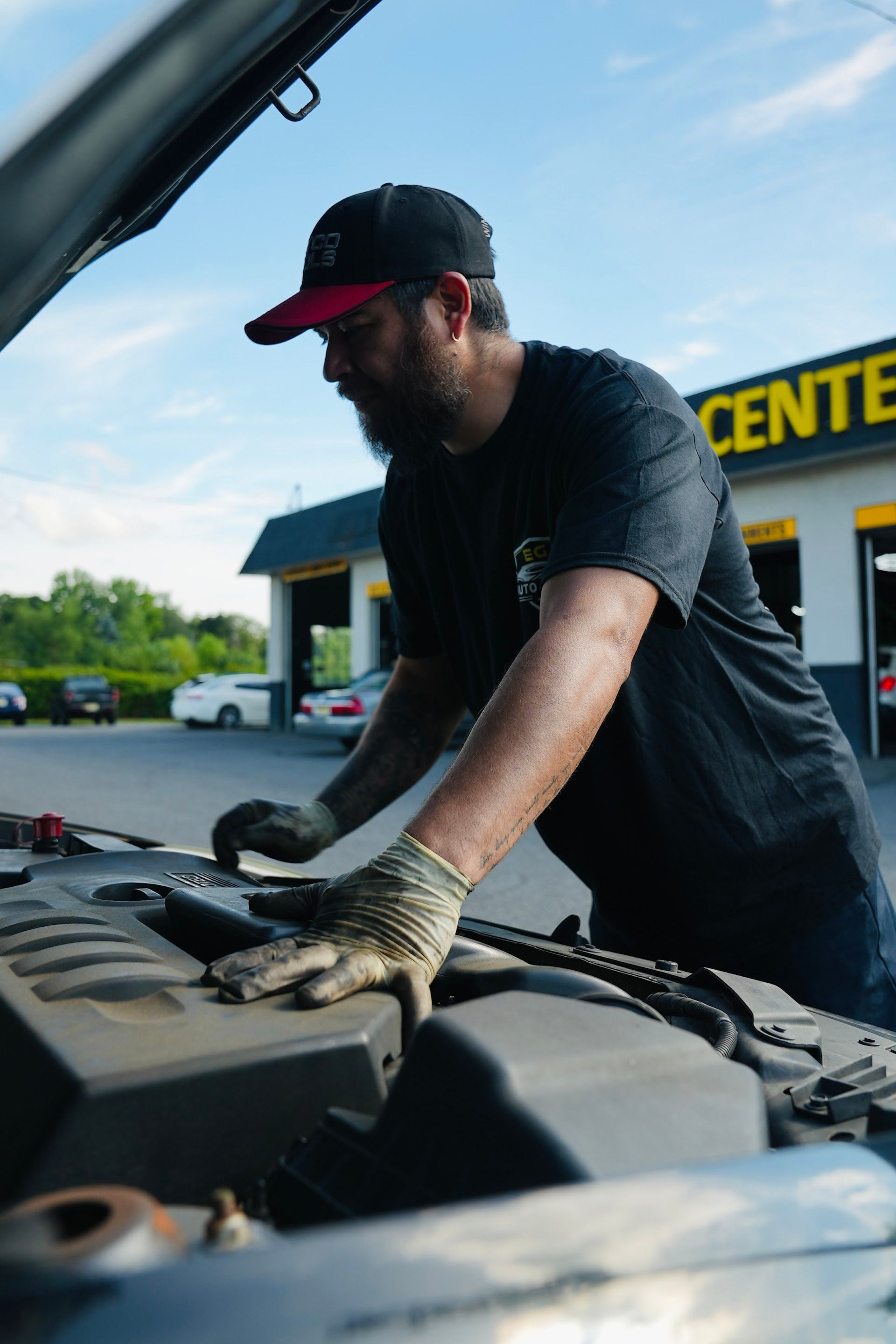 Mechanic inspects car engine with open hood at a repair shop, wearing gloves and a cap. | EG Auto Center