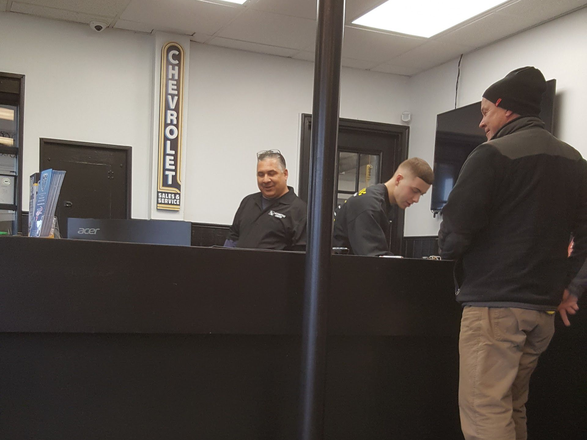 Inside a Chevrolet dealership: people at a service counter. Two employees behind the counter, one smiling. Customer standing nearby. | EG Auto Center