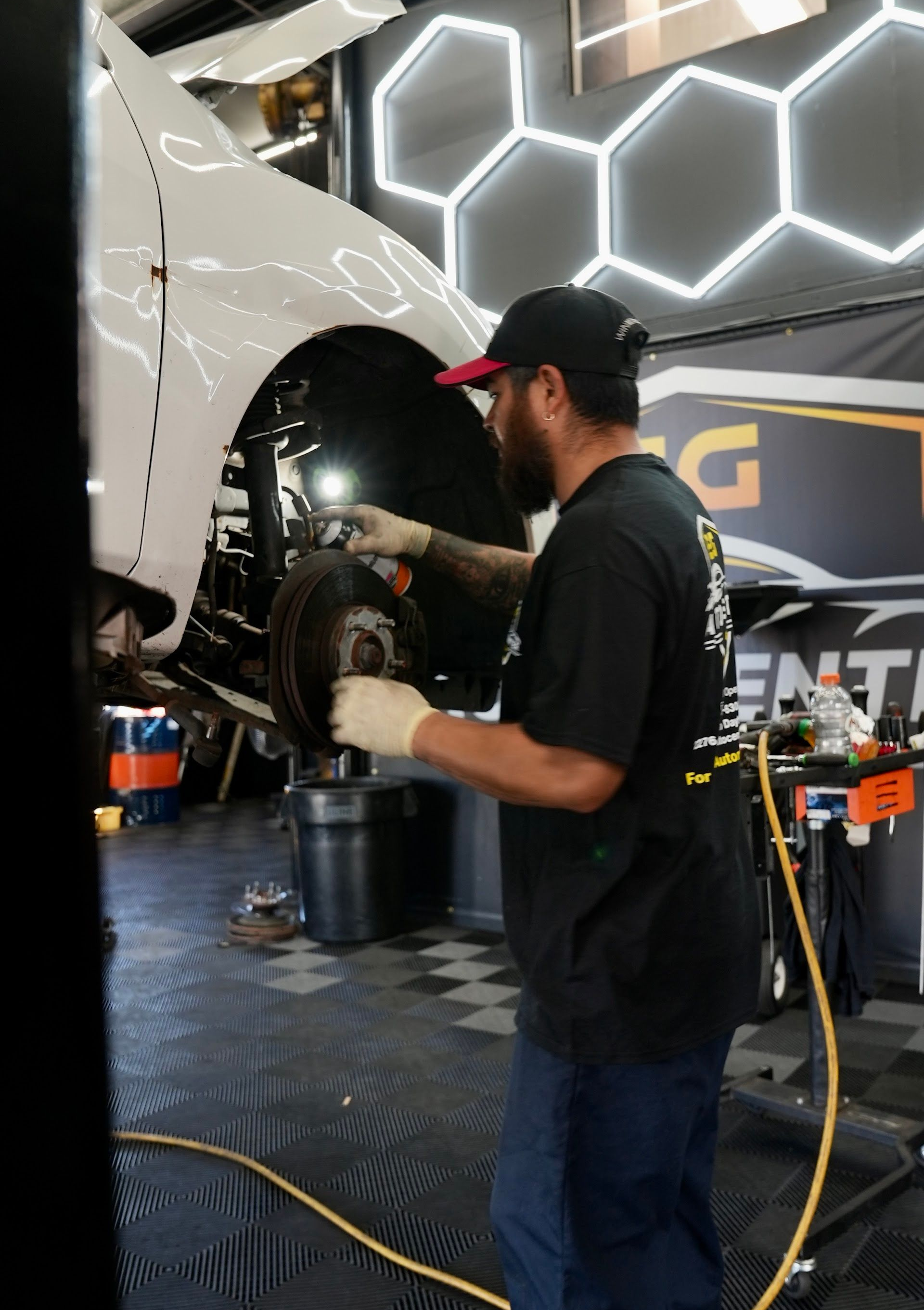 Mechanic working on a car in a garage, using a tool. The car is white, the worker wears a cap and gloves.| EG Auto Center