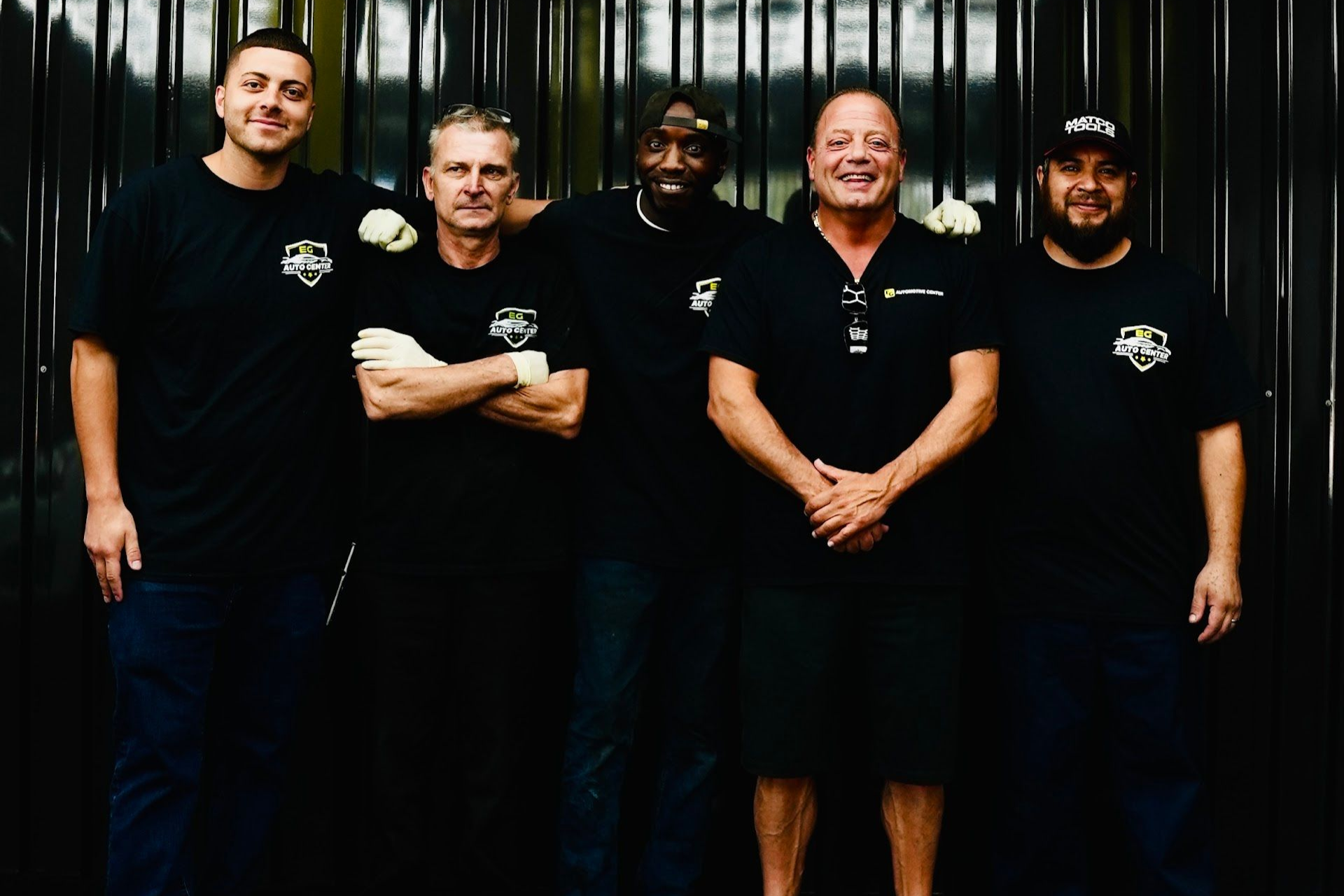 Five people pose in front of a dark backdrop, all wearing black shirts, arms crossed or at sides, smiling. | EG Auto Center