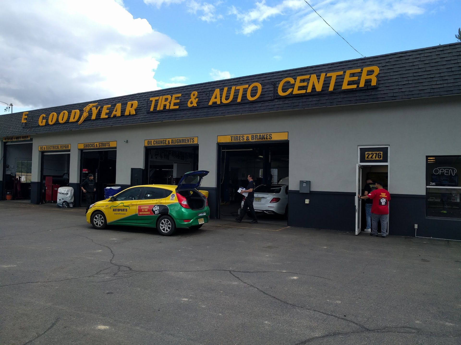 Goodyear Tire & Auto Center exterior. Yellow and green car parked in front of open bay doors. People near doors. | EG Auto Center