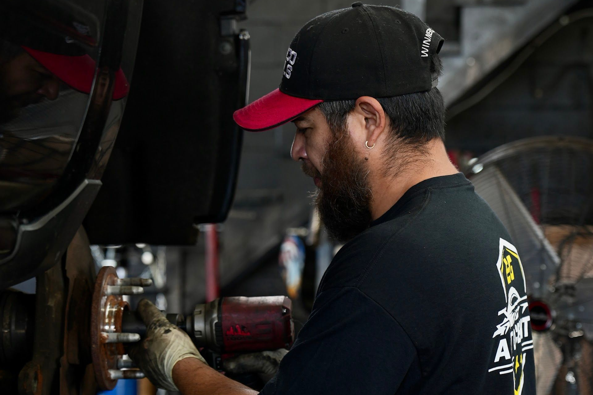 Mechanic with a beard, wearing a black cap and gloves, working on a vehicle in a garage. | EG Auto Center
