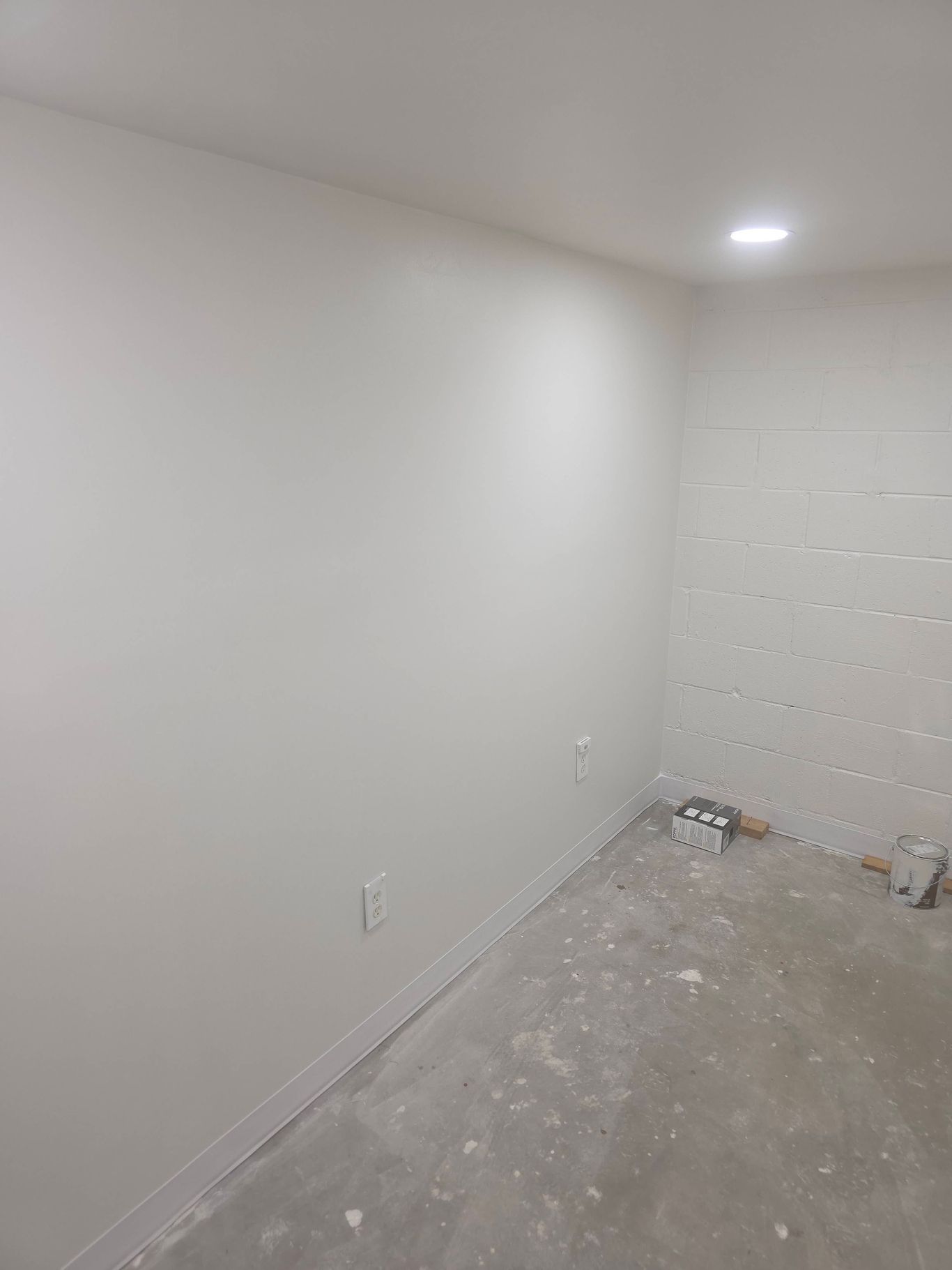 White-painted room corner, unfinished floor, electrical outlet, drywall partially installed, and overhead light.