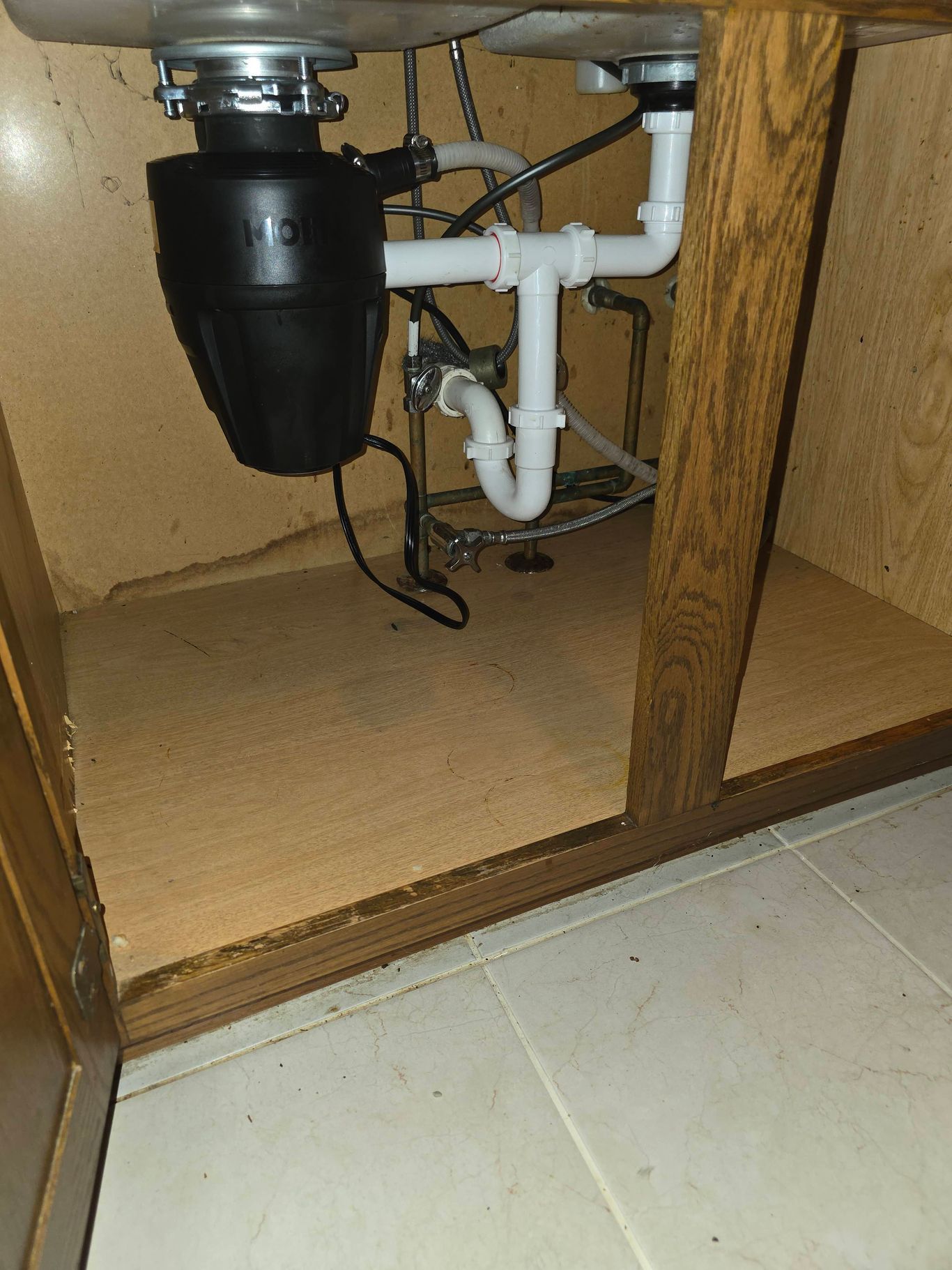 Under-sink view of kitchen cabinet with plumbing, garbage disposal, and light-colored wooden surfaces.