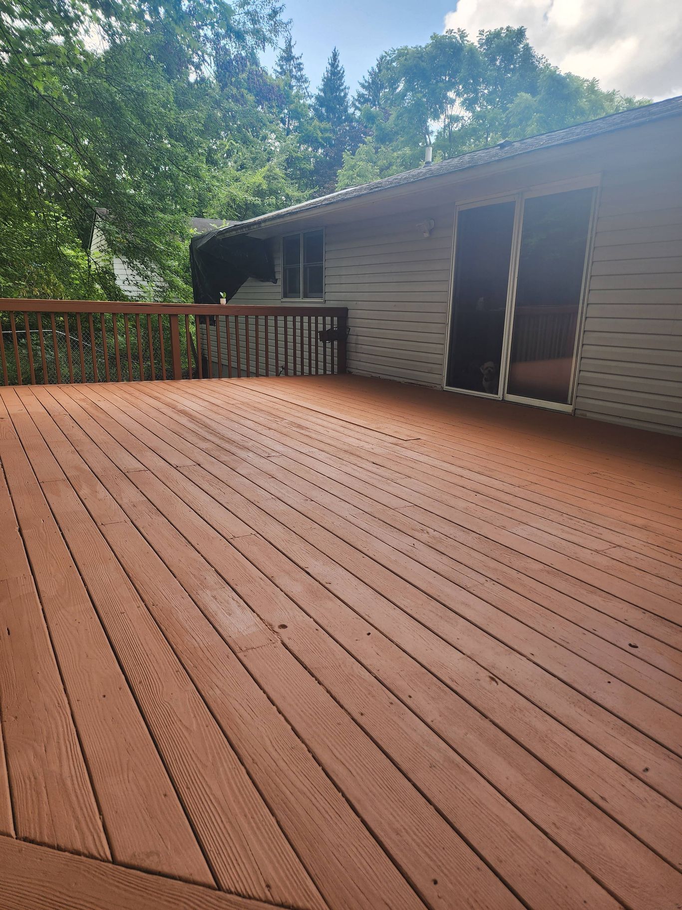 Wooden deck with reddish-brown stain, next to a light-colored house with sliding glass doors, trees in background.