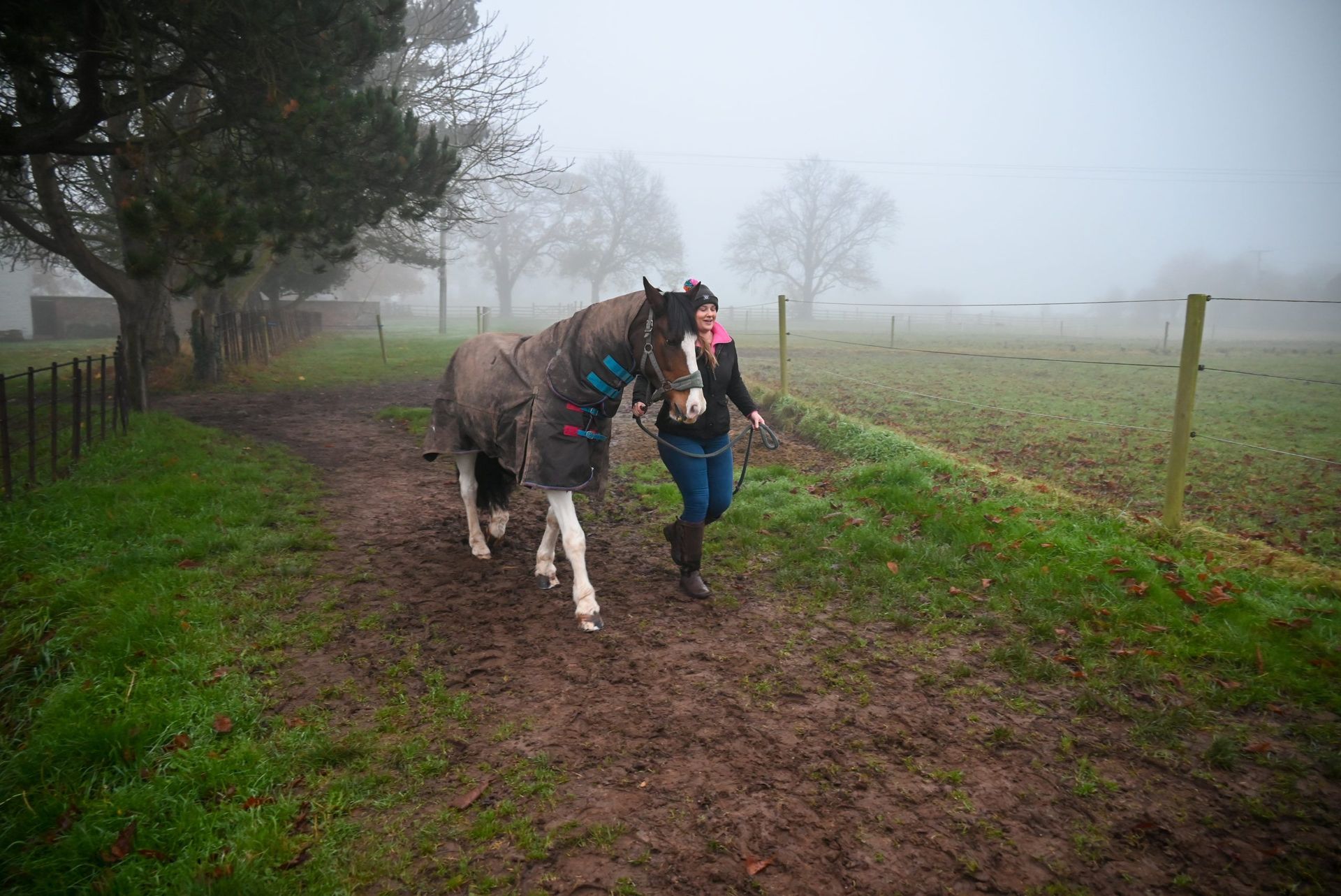 Woman leads a horse wearing a blanket on a muddy path in a field, overcast sky.