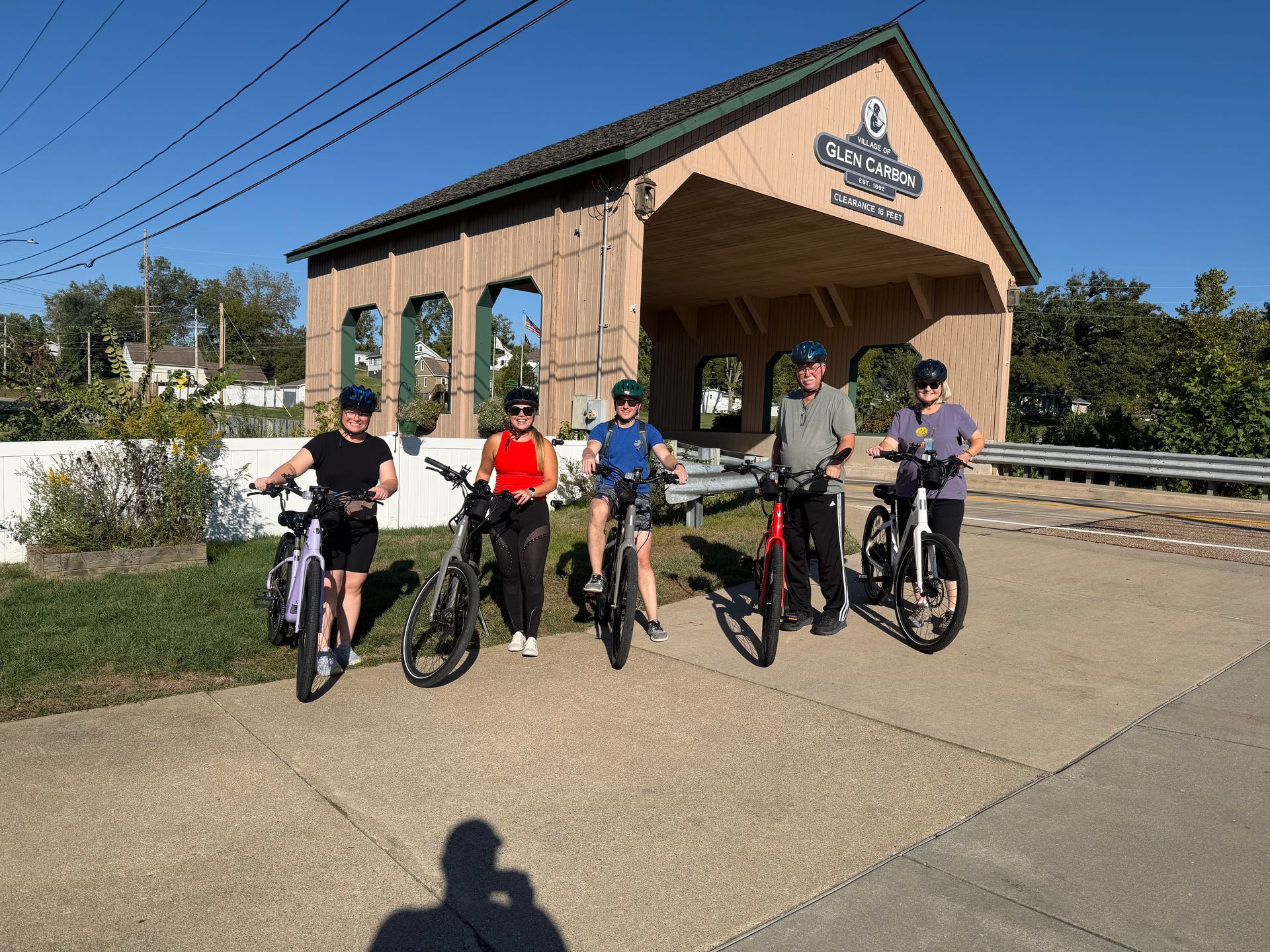 Five cyclists pose with bikes in front of a covered bridge on a sunny day.